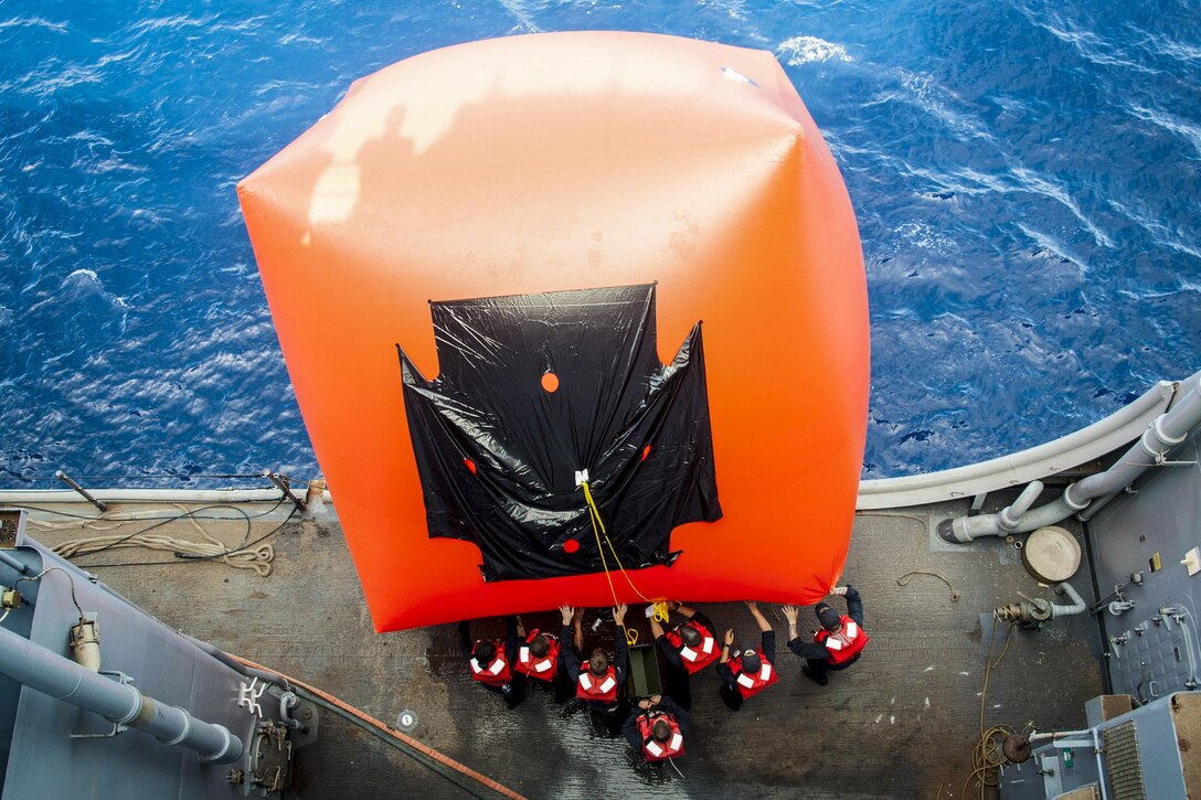 Sailors push a big orange inflatable target off the side of a ship into the water.