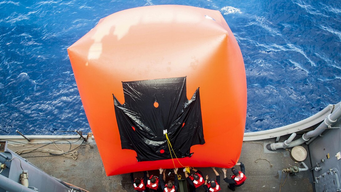 Sailors push a big orange inflatable target off the side of a ship into the water.