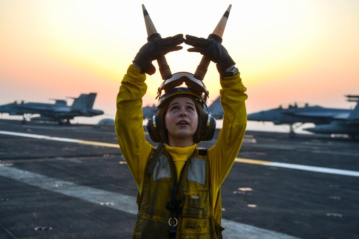 A Sailor signals with her arms on the deck of an aircraft carrier at sunset.