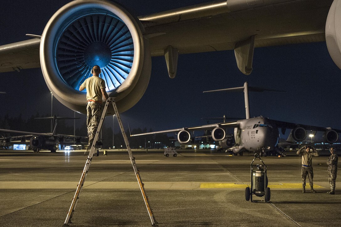 An airman on a ladder on a dark flightline checks an aircraft engine.