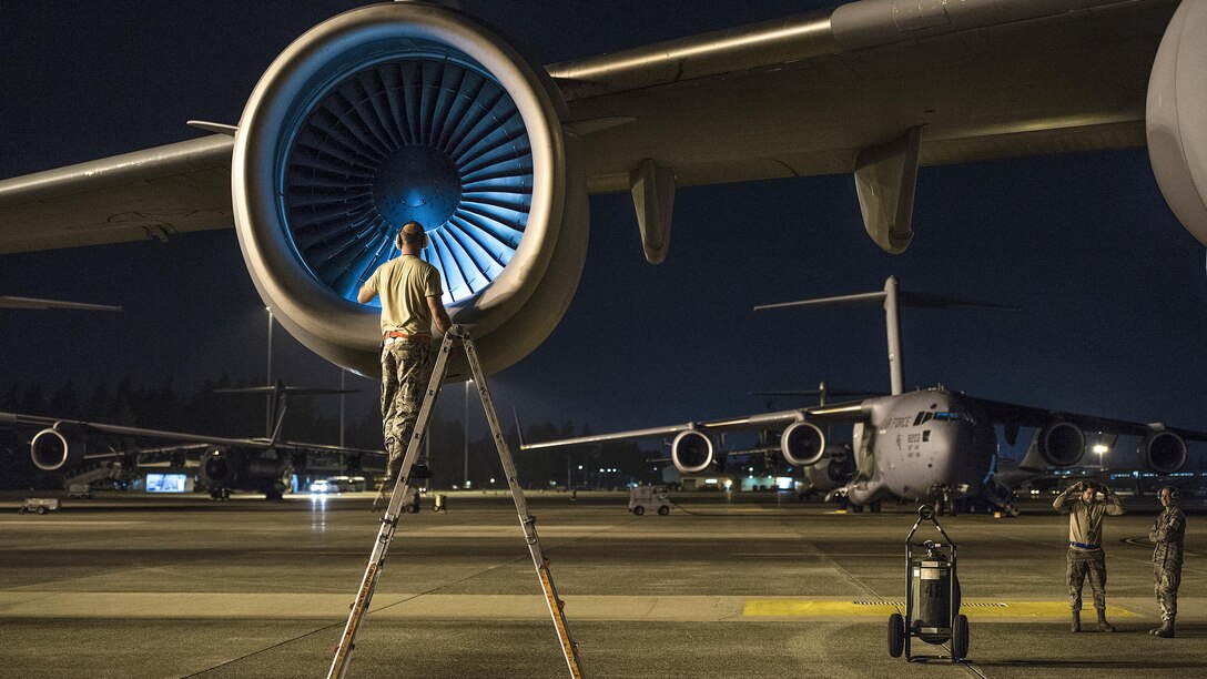 An airman on a ladder on a dark flightline checks an aircraft engine.