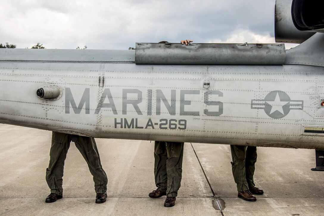 The legs of three Marines are visible behind an aircraft they are inspecting.
