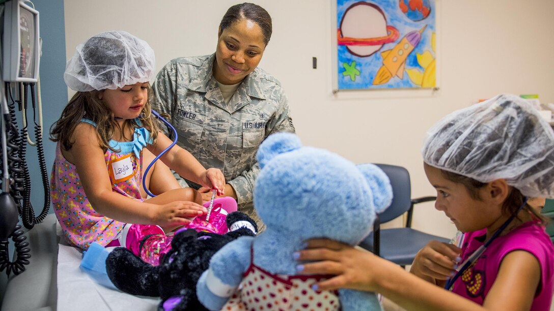 Two girls wearing hair nets tend to teddy bears as an airman watches.