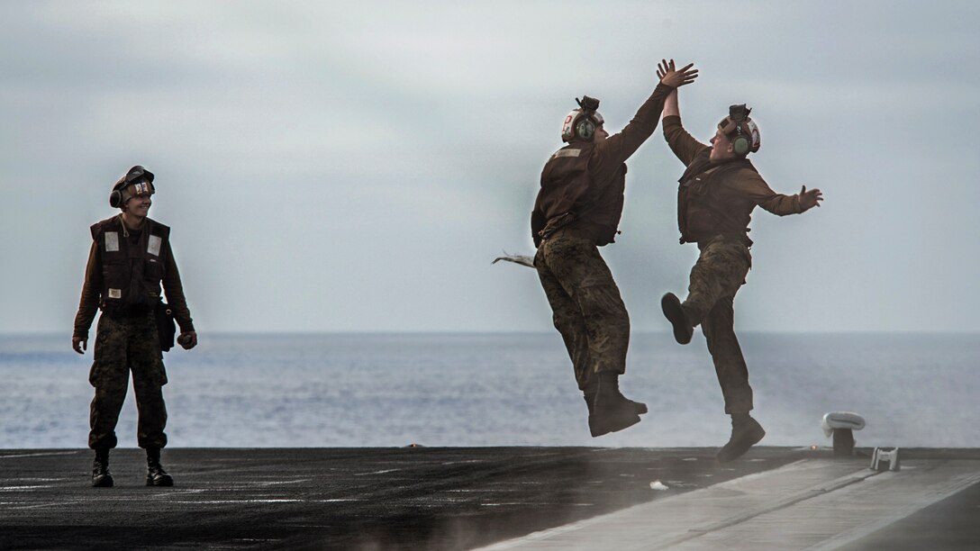 Two Marines jump and high-five each other as another Marine looks on aboard a ship.