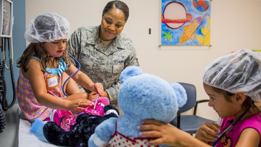 Two girls wearing hairnets tend to teddy bears as an airman watches.