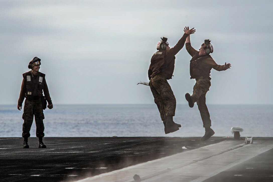 Two Marines jump and high-five each other as another Marine looks on aboard a ship.