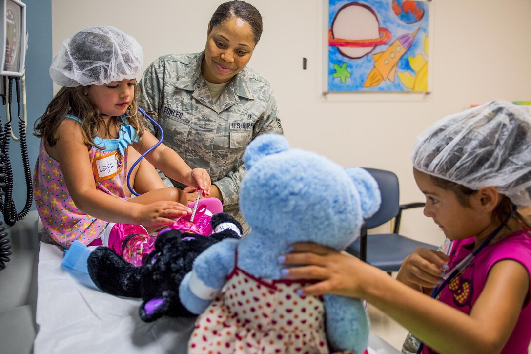 Two girls wearing hairnets tend to teddy bears as an airman watches.