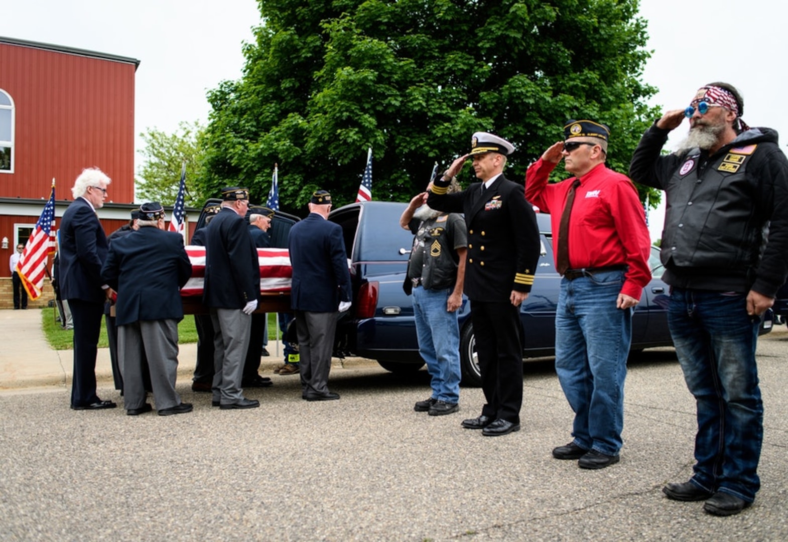 Pallbearers at rear of frame, unloading flag-draped casket from hearse; at right, civilians and Navy captain in class A uniform stand at attention.