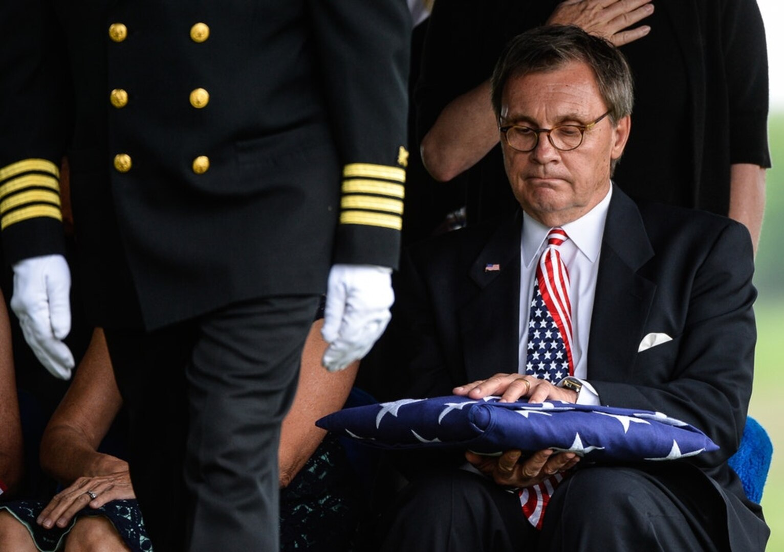 Seated civilian in suit, somberly holding folded US flag; at left, Navy captain in class A uniform moving toward viewer.