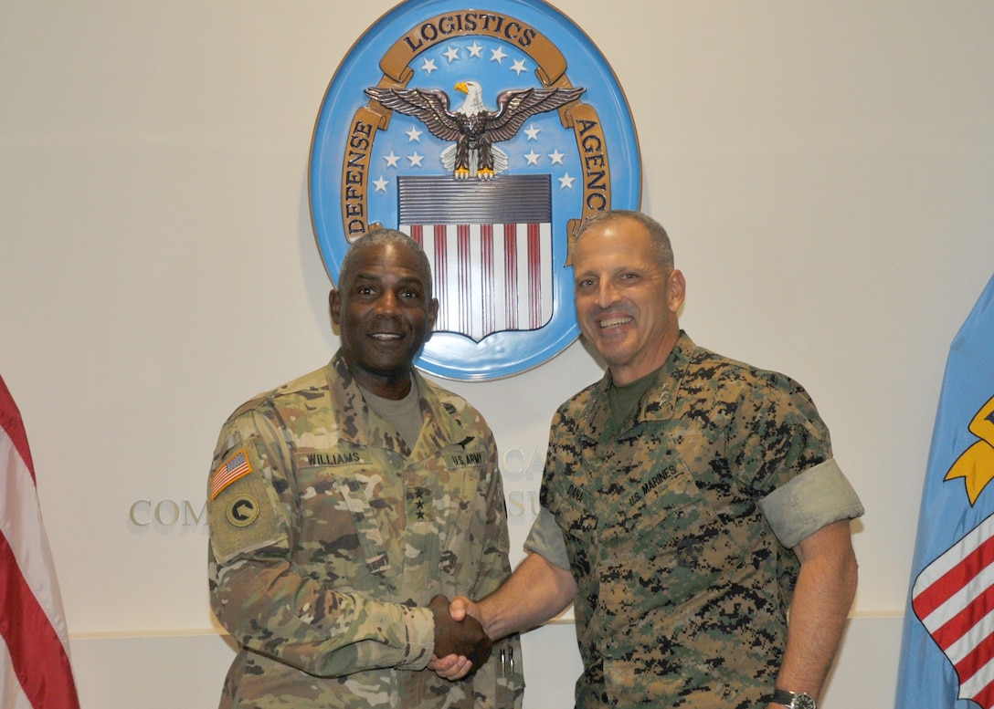 Facing viewer, Army general and Marine general in fatigues, shaking hands; DLA seal in background and flags on either side.