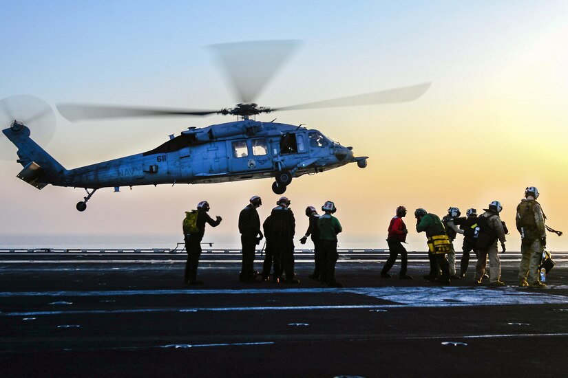 Troop stand on a ship's flight deck as a helicopter comes in for a landing.