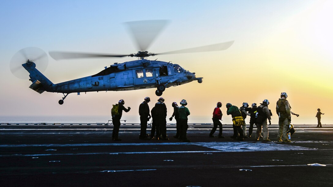 Troop stand on a ship's flight deck as a helicopter comes in for a landing.