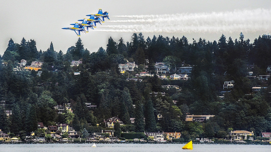 Four aircraft fly in formation over a lake and lakeside houses.