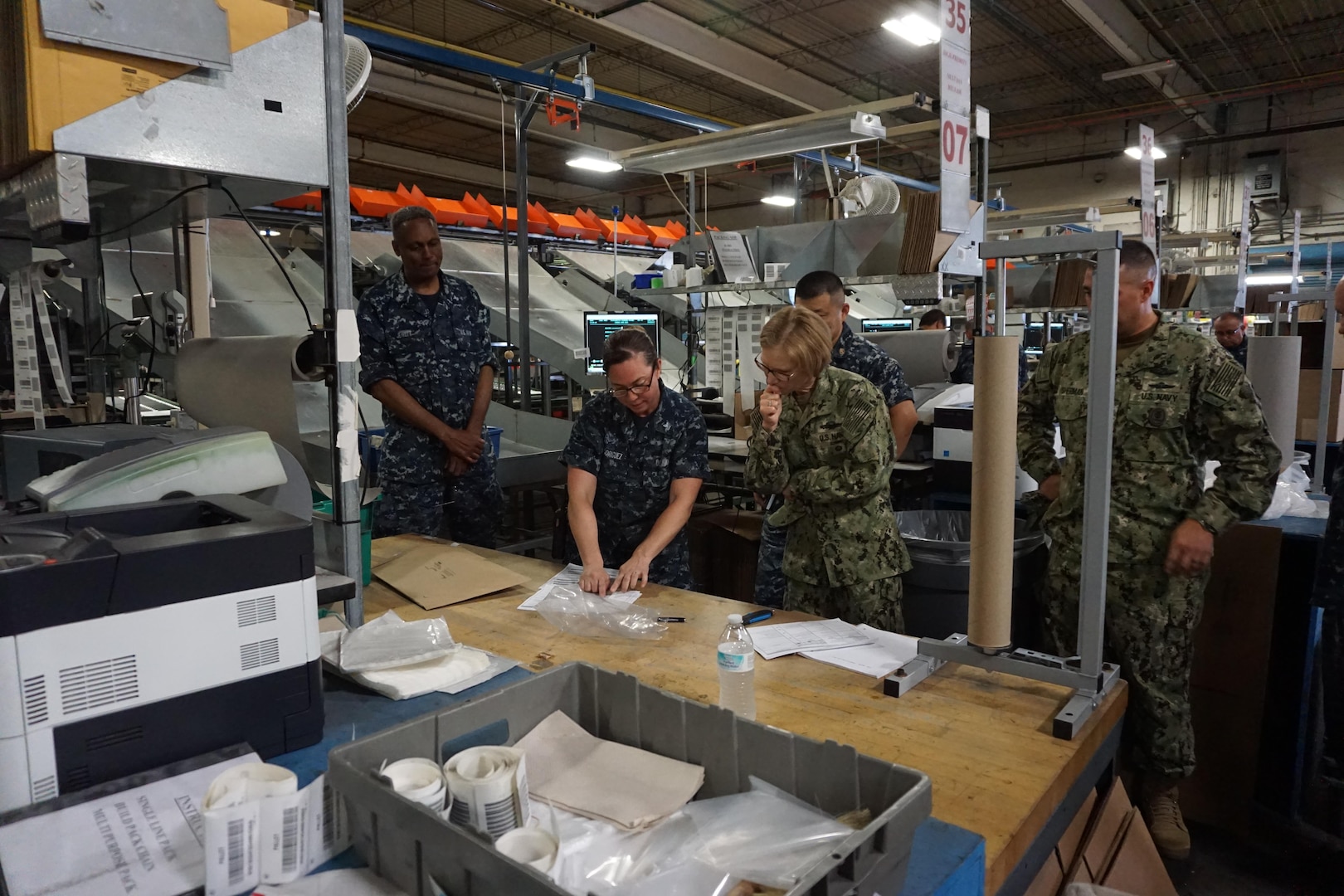 A group of people gathered around a worktable in a distribution warehouse observe someone using a shipping label and other shipping supplies in a demonstration.