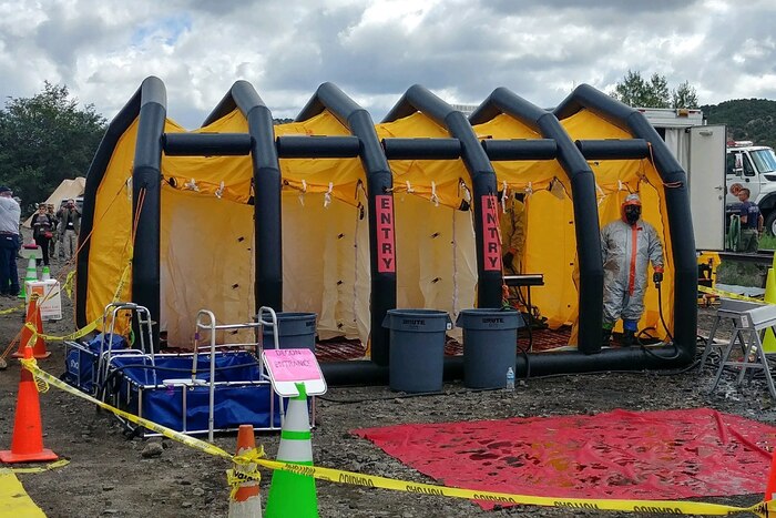 A man wearing full decontamination gear stands in a large yellow and black mass decontamination shower.