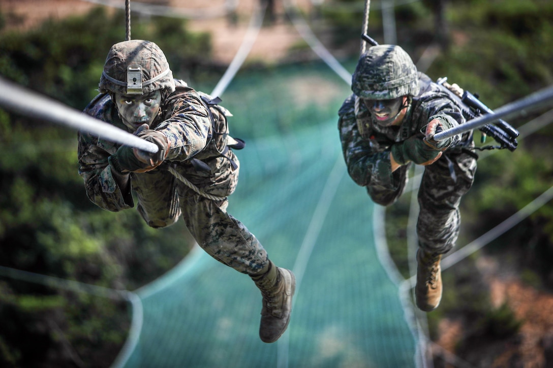 Two service members pull themselves across rope obstacles.