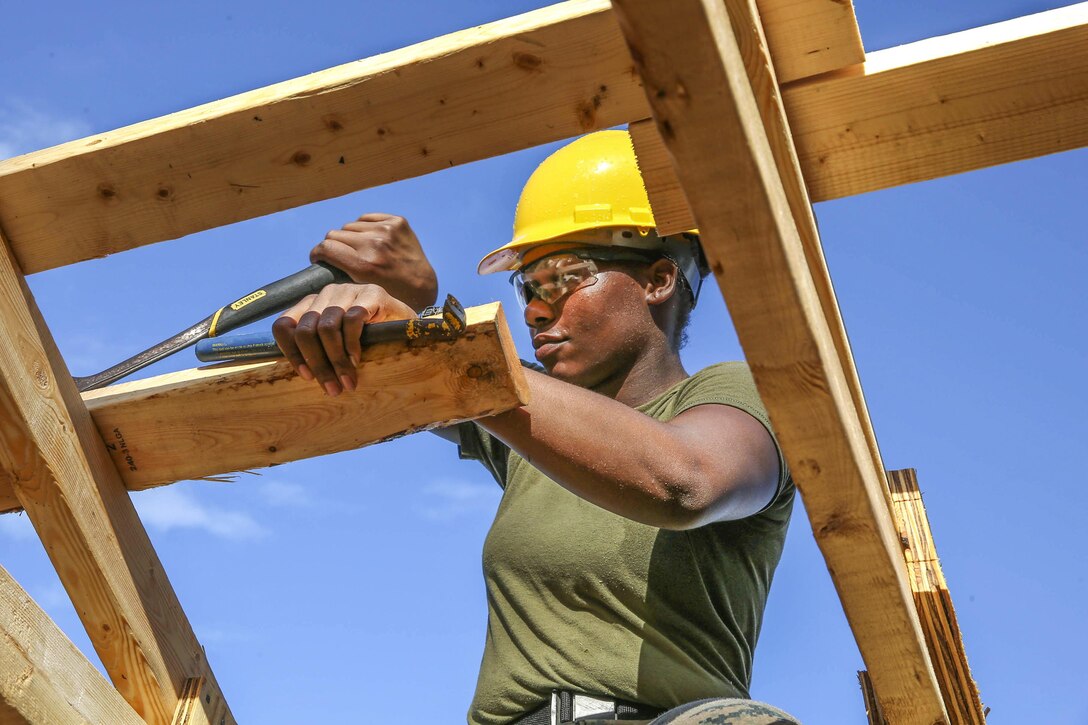 A Marine wearing a hard hat uses tools to help build a wooden structure.
