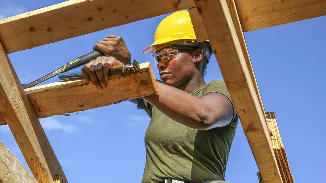 A Marine wearing a hard hat uses tools to help build a wooden structure.