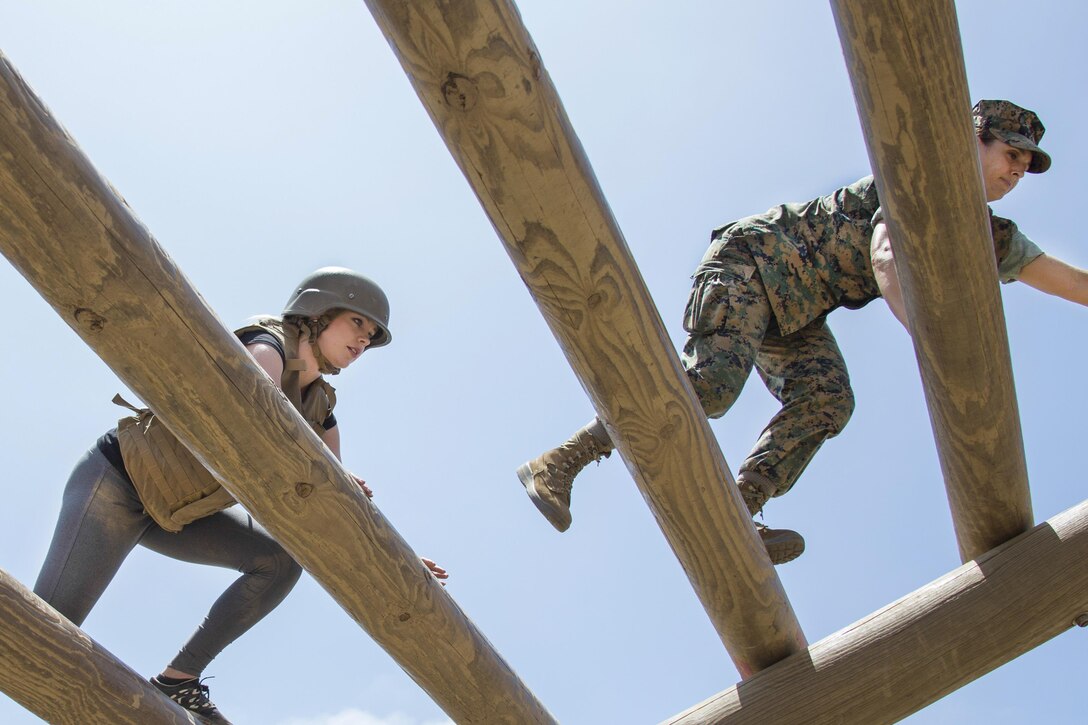 A Marine climbs a wooden ladder obstacle as a reporter follows behind.