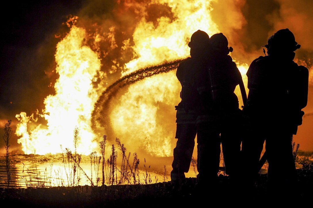 Three firefighters, shown in silhouette, spray water on a fire.