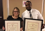 Facing viewer, woman and man in business attire posing with framed awards.
