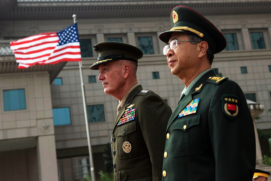 Marine Corps Gen. Joe Dunford walks next to his Chinese counterpart with an American flag in the background.