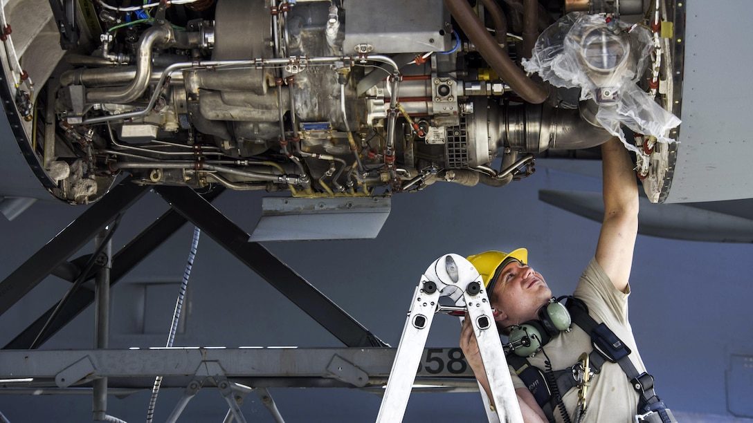 An airman on a ladder reaches up to an aircraft engine.