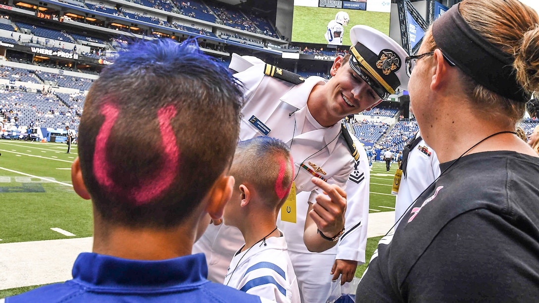 A sailor points at a horseshoe logo painted on a boy's head a boy with a similar logo looks on.