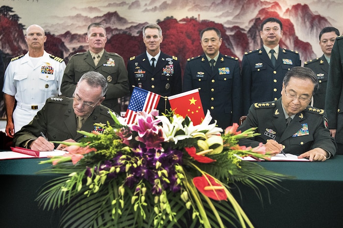 Two military leaders sign documents at a table with a group of people behind them.
