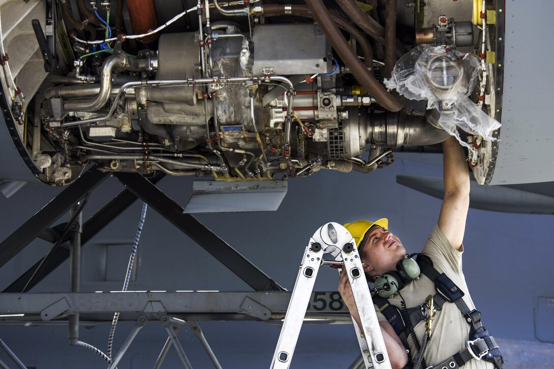 An airman on a ladder reaches up to an aircraft engine.