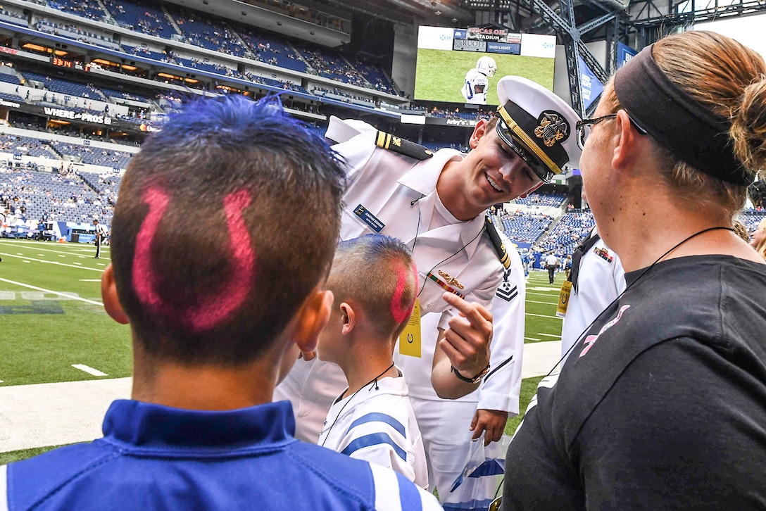 A sailor points at a horseshoe logo painted on a boy's head a boy with a similar logo looks on.
