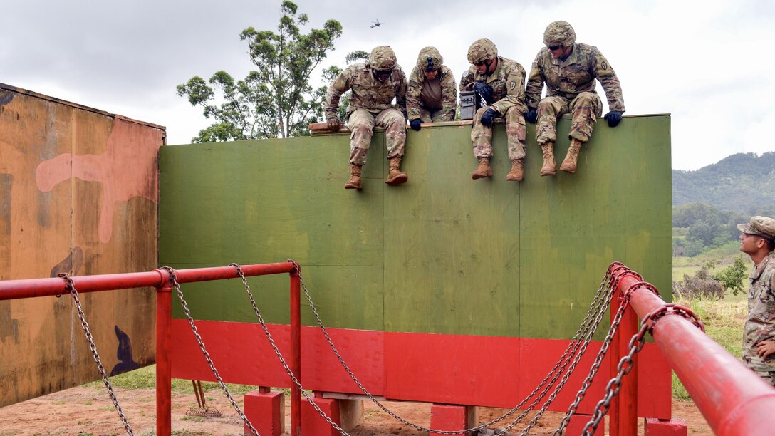 Four soldiers confer while sitting on a high green wall.
