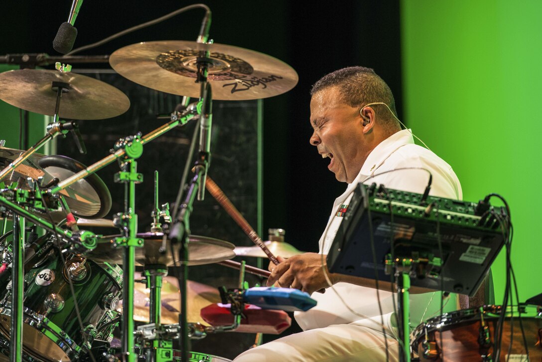 This image shows a sailor enjoying himself as he plays drums in front of a green wall.