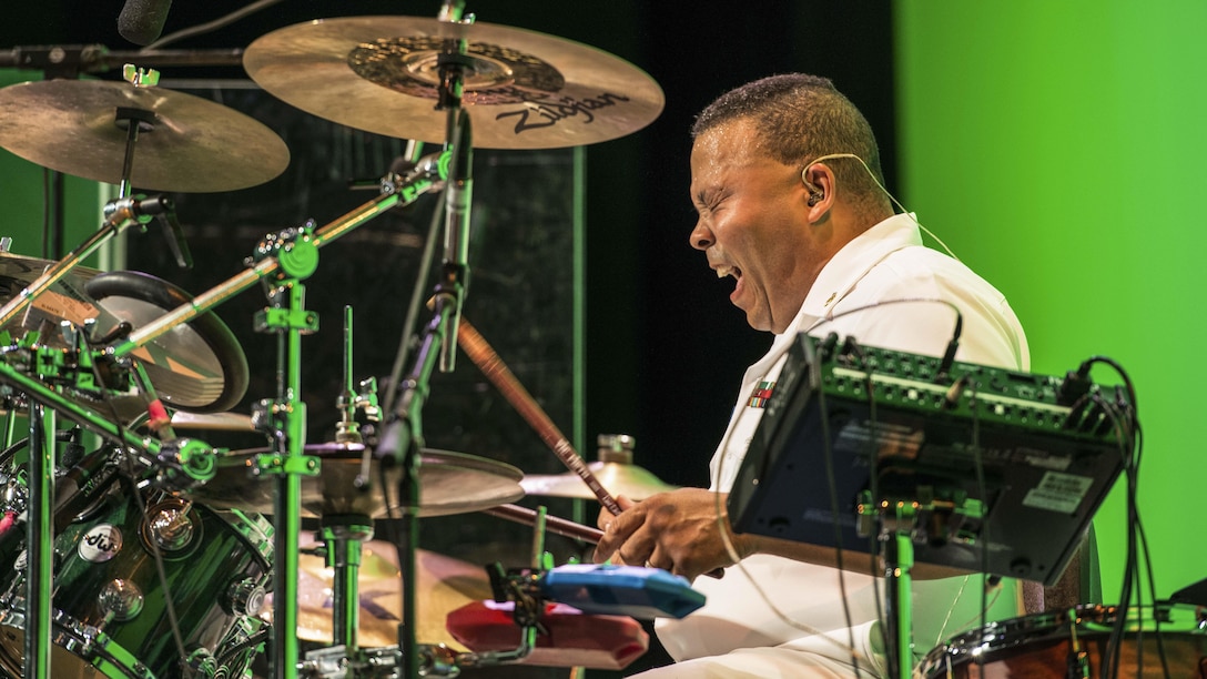 This image shows a sailor enjoying himself as he plays drums in front of a green wall.