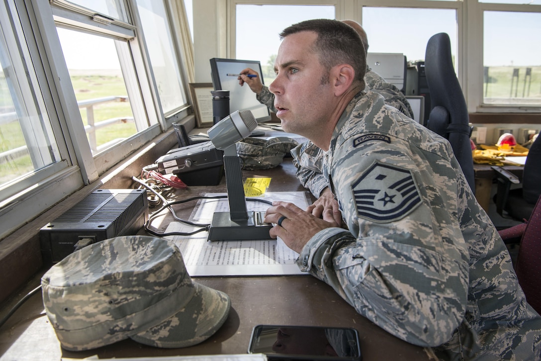 An airman sits at a table watching out a window and speaking into a microphone.