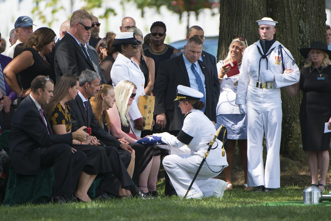 A sailor kneels and presents a folded U.S. flag to a woman seated during a graveside service.