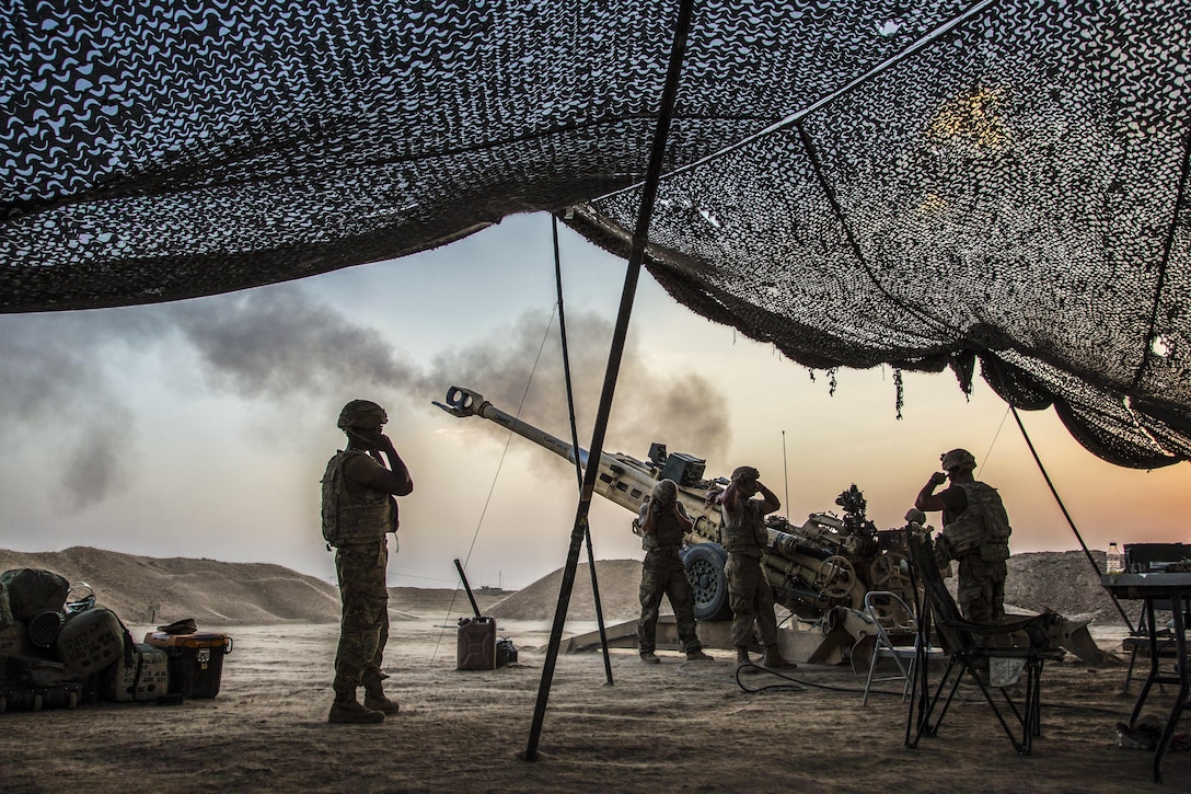 Soldiers standing near overhead netting cover their ears as a howitzer is fired.