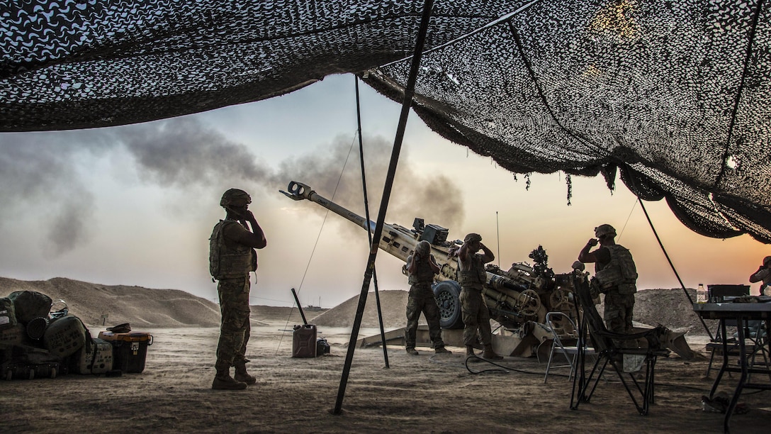 Soldiers standing near overhead netting cover their ears as a howitzer is fired.