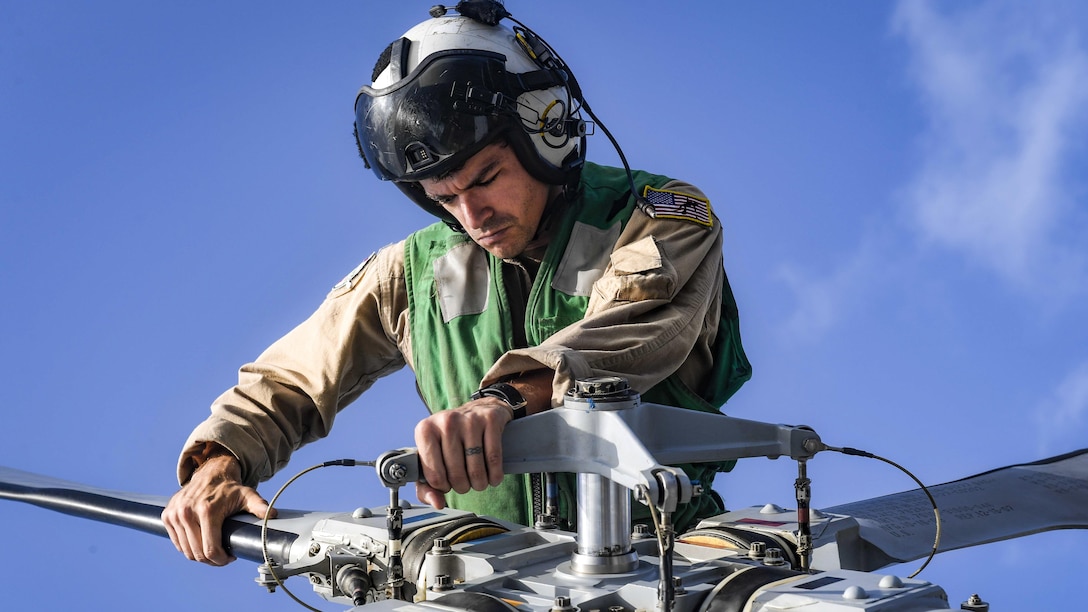 A sailor works on a rotor blade while sitting on top of a helicopter.