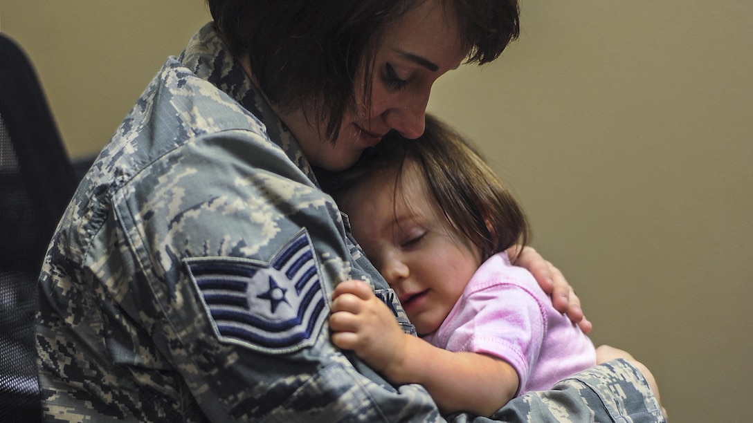 A softly smiling mother in an Air Force uniform holds her softly smiling baby girl.