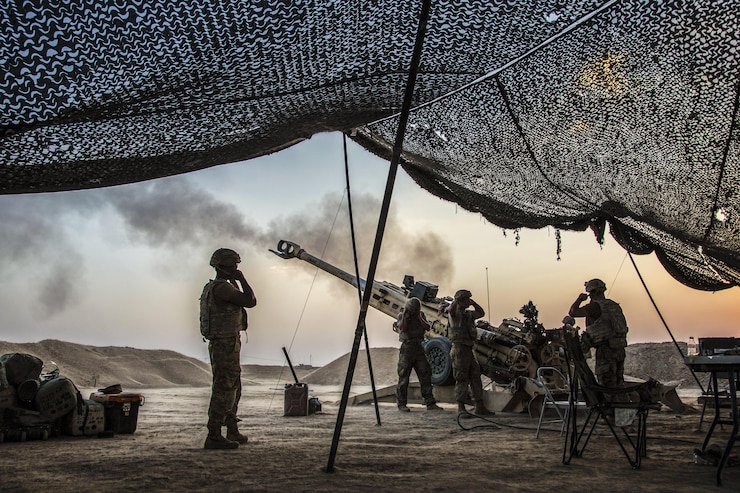 Soldiers standing near overhead netting cover their ears as a howitzer is fired.