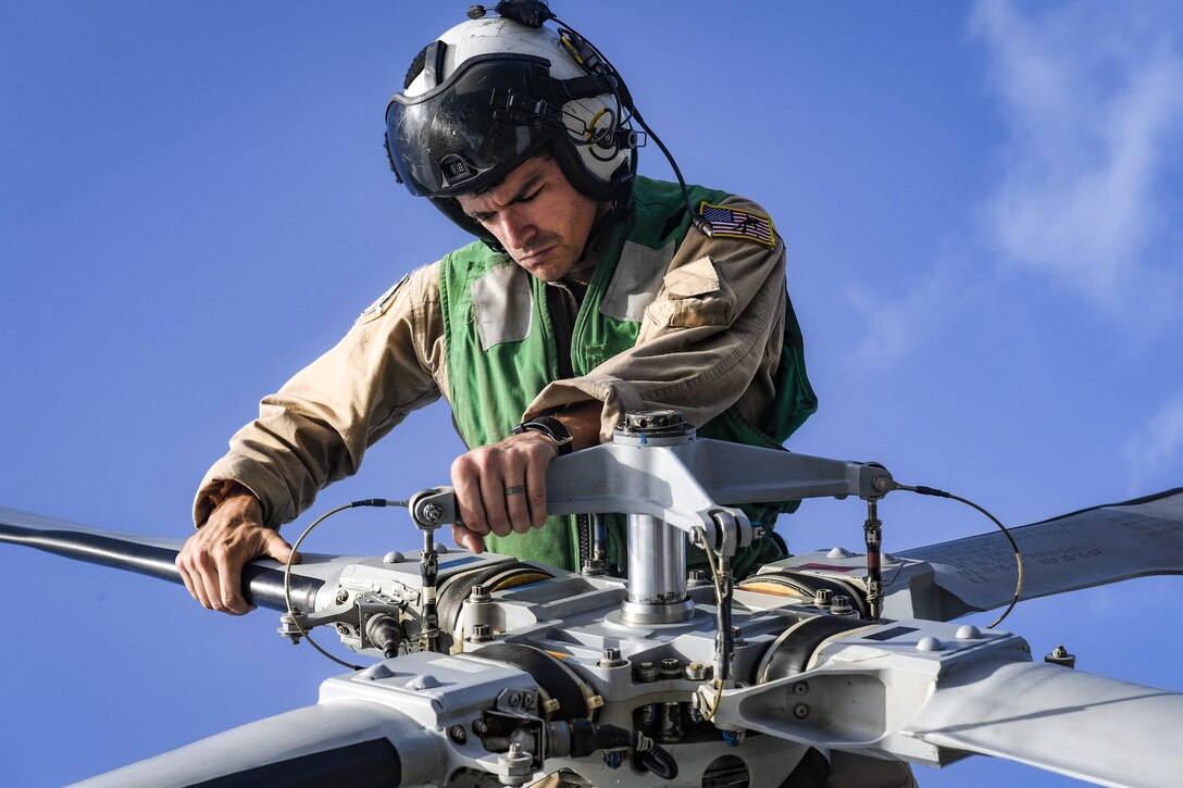 A sailor works on a rotor blade while sitting on top of a helicopter.