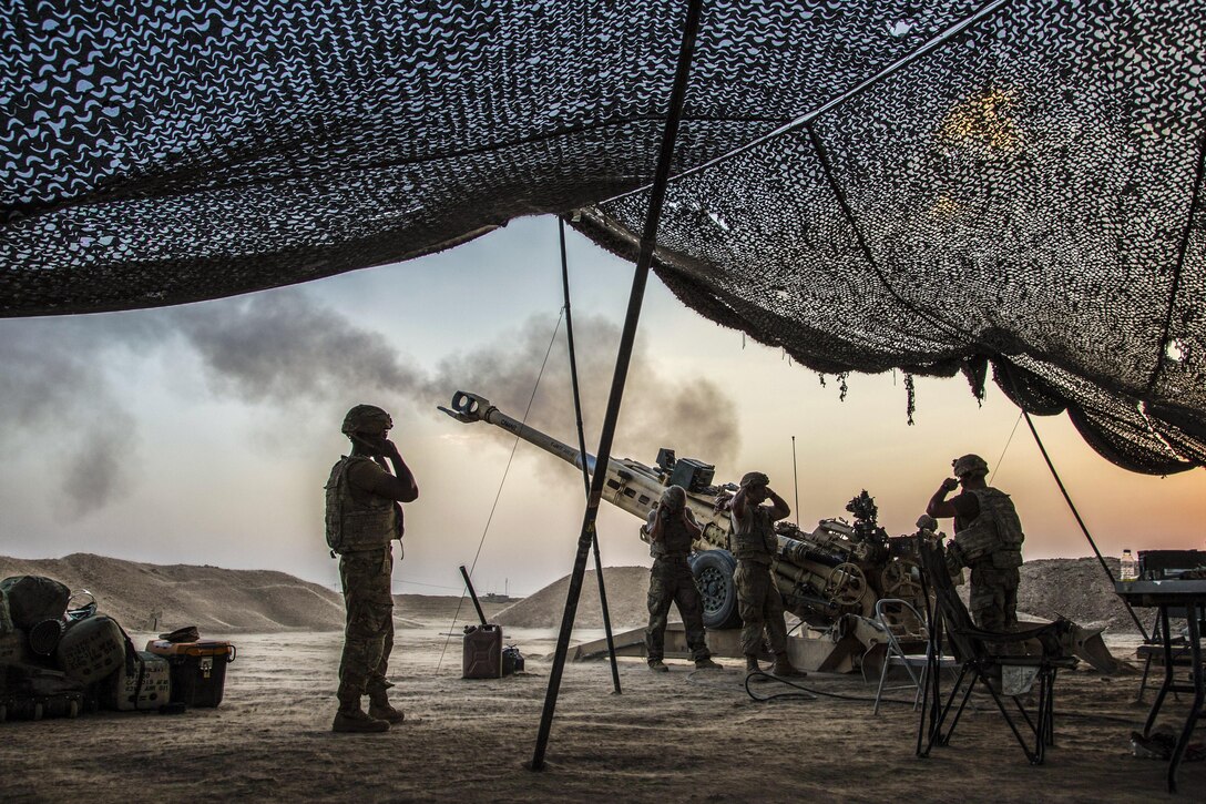 Soldiers standing near overhead netting cover their ears as a howitzer is fired.