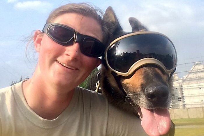 A smiling woman's face next to a dog's face, the dog is wearing goggles.