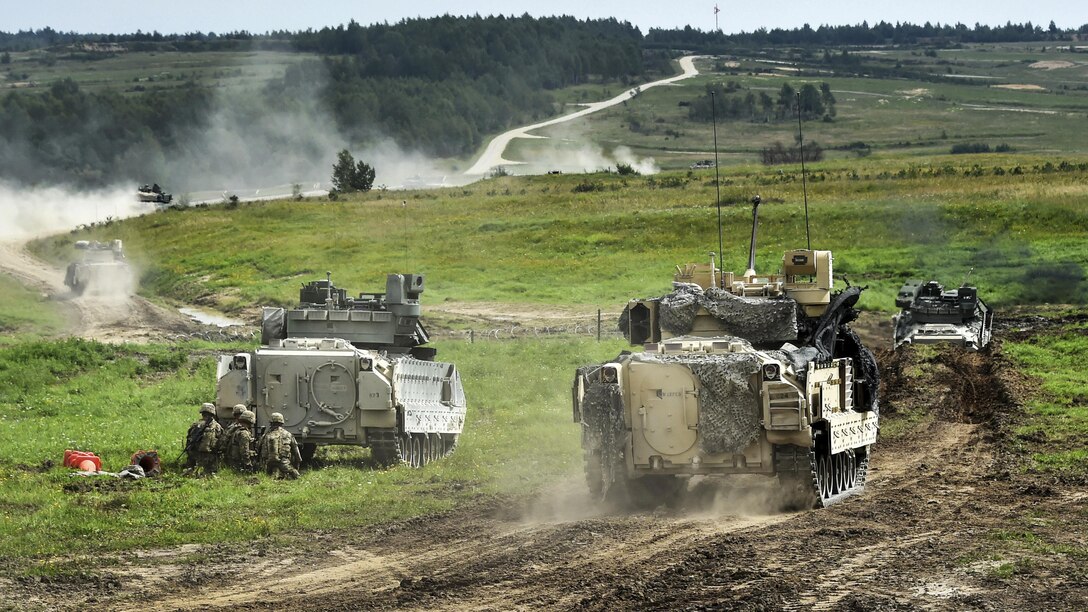 Soldiers in Abrams tanks and Bradley vehicles participate in a live-fire exercise in a green field.