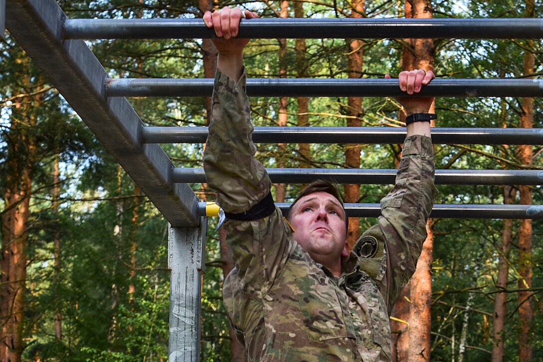A soldier crosses a monkey bar obstacle in a wooded area.