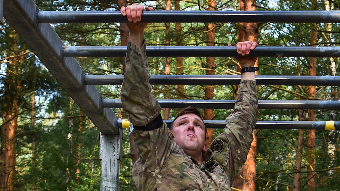 A soldier crosses a monkey bar obstacle in a wooded area.