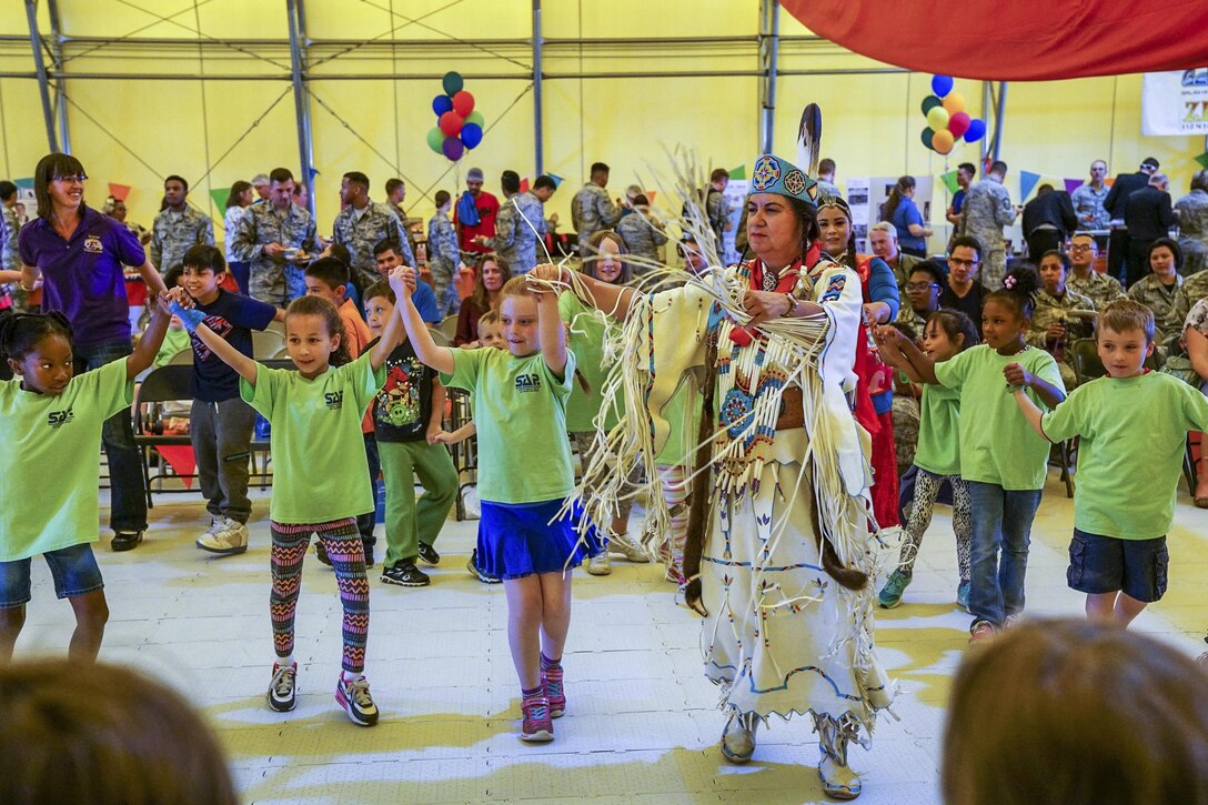 Children hold hands and dance with a person in Native American garb as troops look on and mingle.