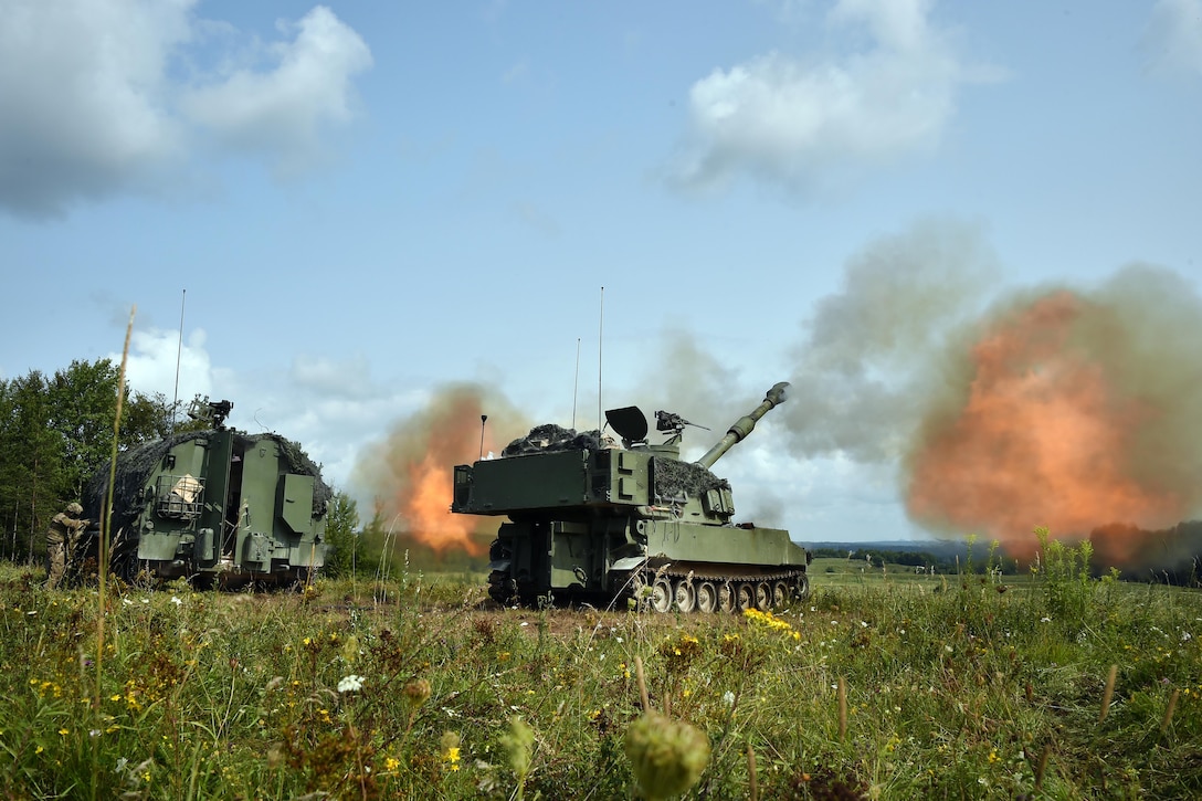 Soldiers in artillery vehicles fire weapons in a green field, creating orange-colored smoke.