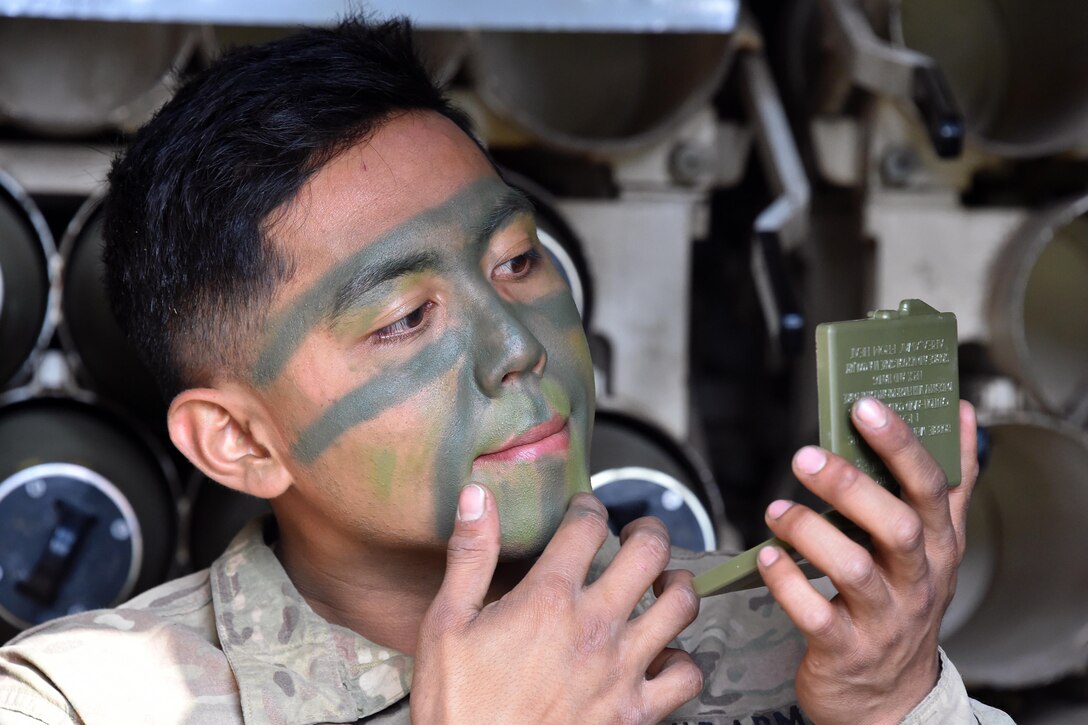 A soldier applies camouflage paint to his face while holding a mirror.